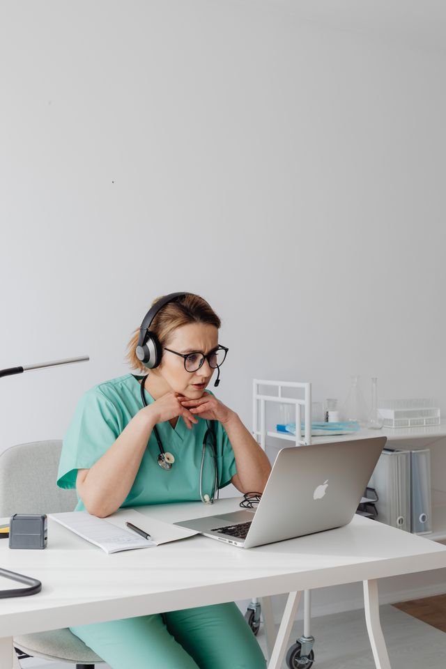 A medical professional focuses intently on a laptop while wearing teal scrubs and headphones.